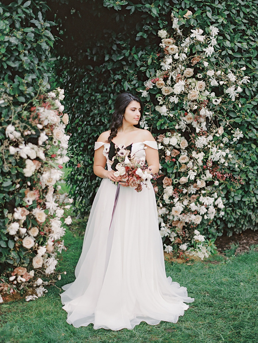 Folk Art Flowers Chateau Lill Spring Wedding Mauve Black and White Flowers A bride holding a bouquet stands in front of a manicured hedge "doorway" to the vineyard at Chateau Lill