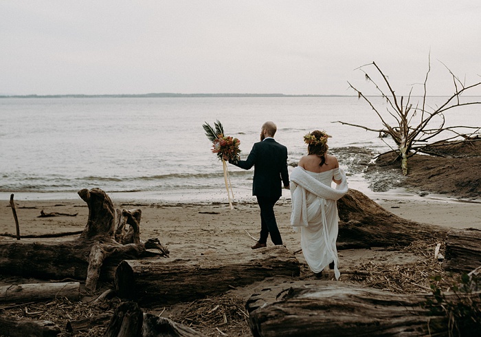 The groom (holding the bouquet) and bride walk towards the beach