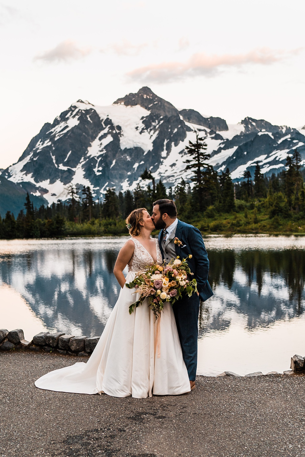 Mountain-Wedding-with-Vibrant-Summer-Blooms-Sunset-Lake-Kiss A couple kisses in front of a lake with a large mountain in the background