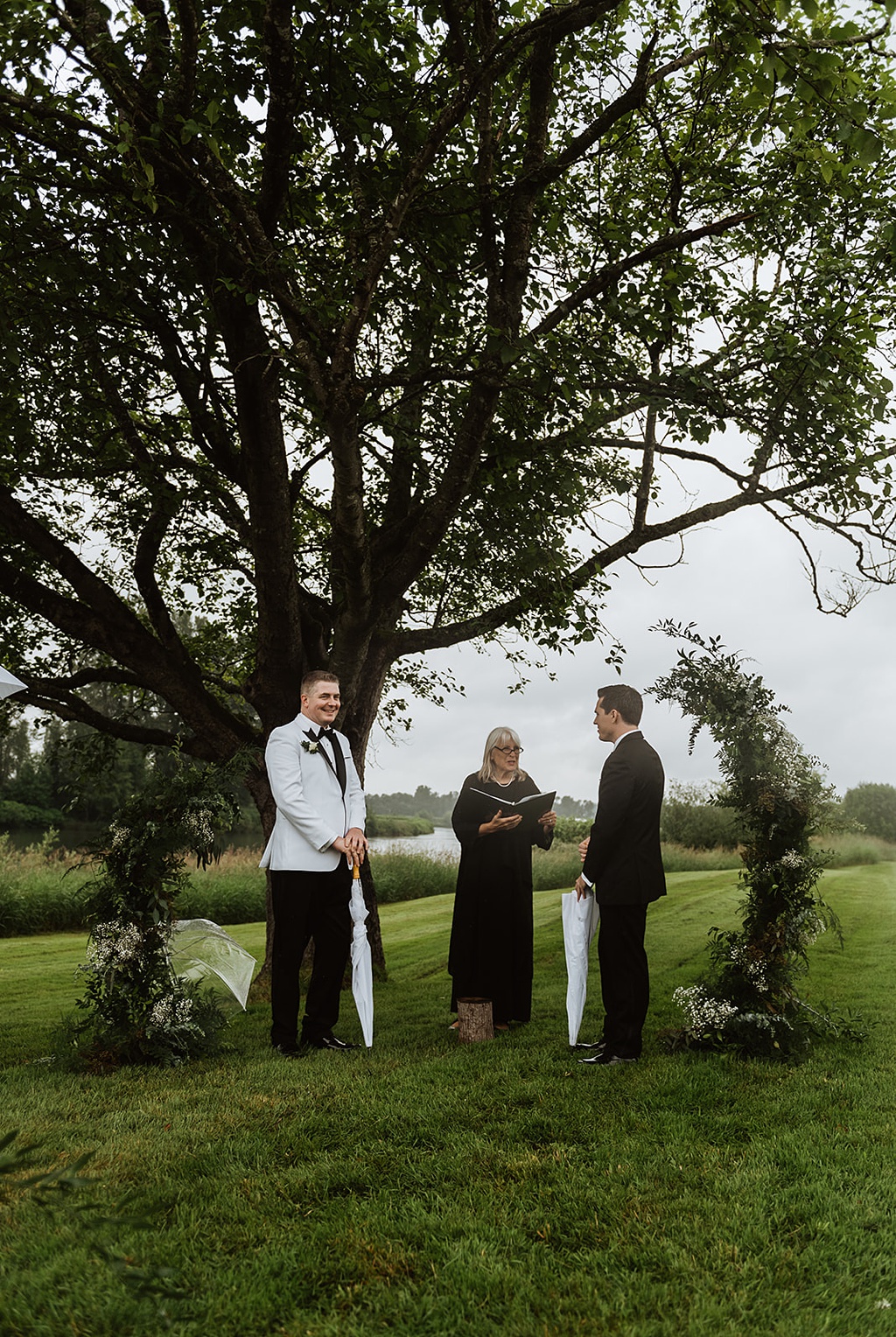 Two grooms standing at a large tree and floral arch for their wedding ceremony